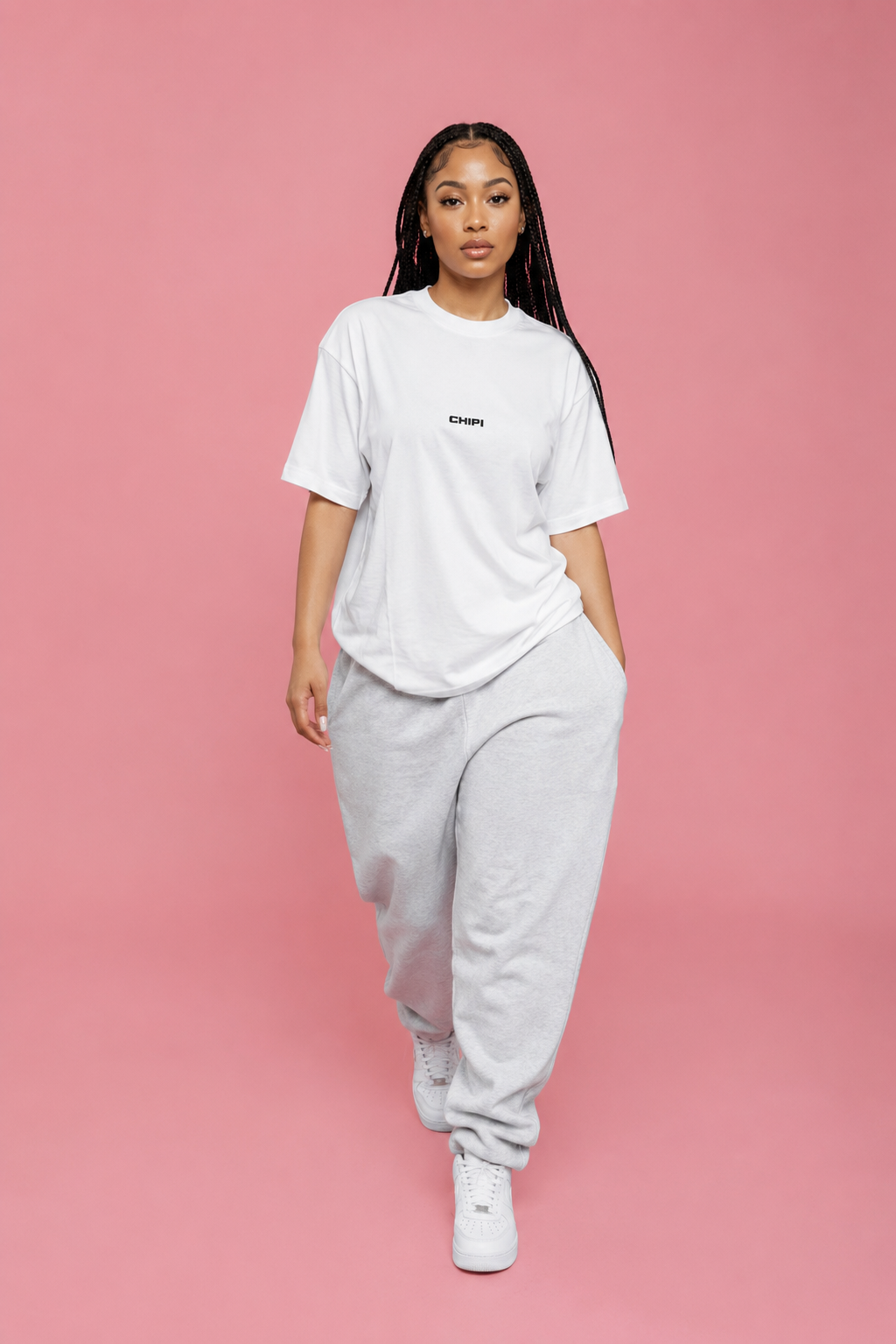 A woman modeling a white crewneck t-shirt with a small black logo on the chest, paired with light gray sweatpants and white sneakers, set against a solid pink studio background.